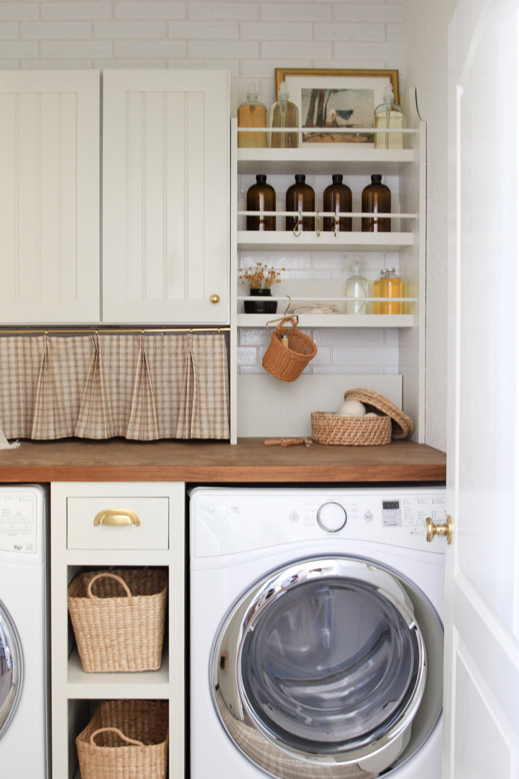 Laundry room, cabinetry mixed with open shelving for storage. Styled with functional glass bottles, pleated curtain, and woven baskets.