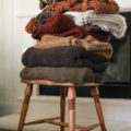 An assortment of cozy blankets displayed atop a vintage wooden chair. Blog post is about cozy fall blankets and includes links to a curated collection by Iris Nacole.