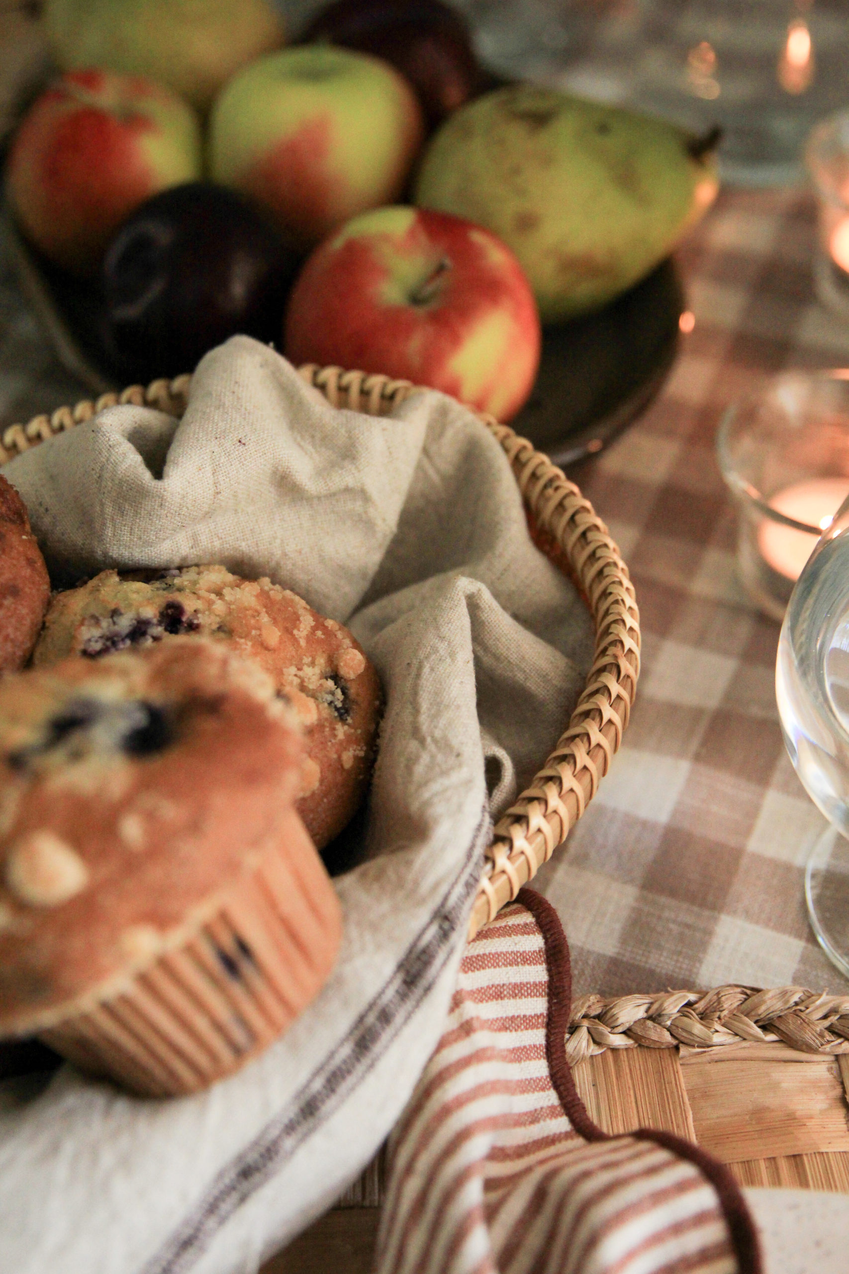 Table setting showcasing a woven basket holding assorted muffins, on a gingham tablecloth. Plate of fruit in background.