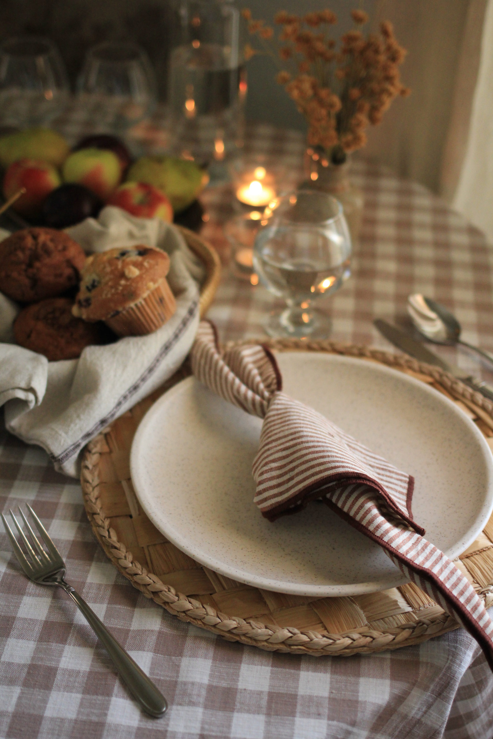 Table setting, featuring a gingham tablecloth, woven charger, speckled pottery plate, with a striped cloth napkin displayed atop the plate.  Recycled glass glasswaer, and vintage style silverware.